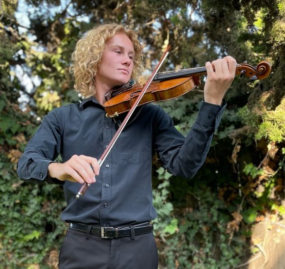 Young man with blonde, curly, shoulder-length hair wearing a black dress shirt and black pants holding a violin and bow in playing position.